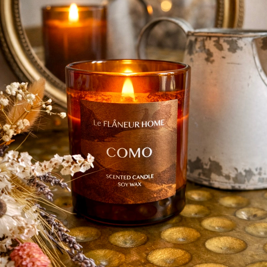 Scented candle labeled 'Le Flèneur Home Como' on a decorative surface with flowers and another candle in the background.