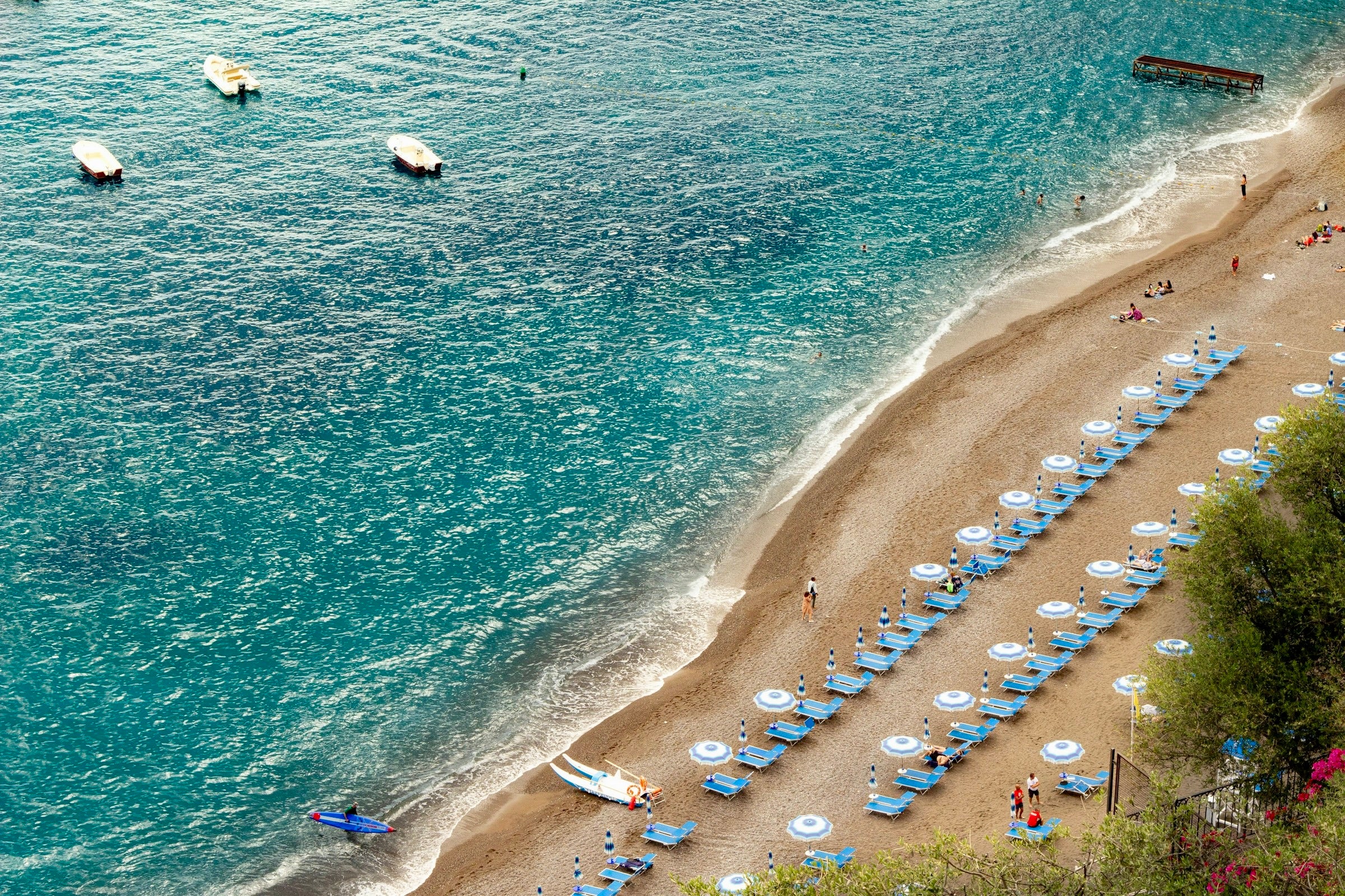 Positano beach with blue lounge chairs and umbrellas along a sandy shore with clear blue water.