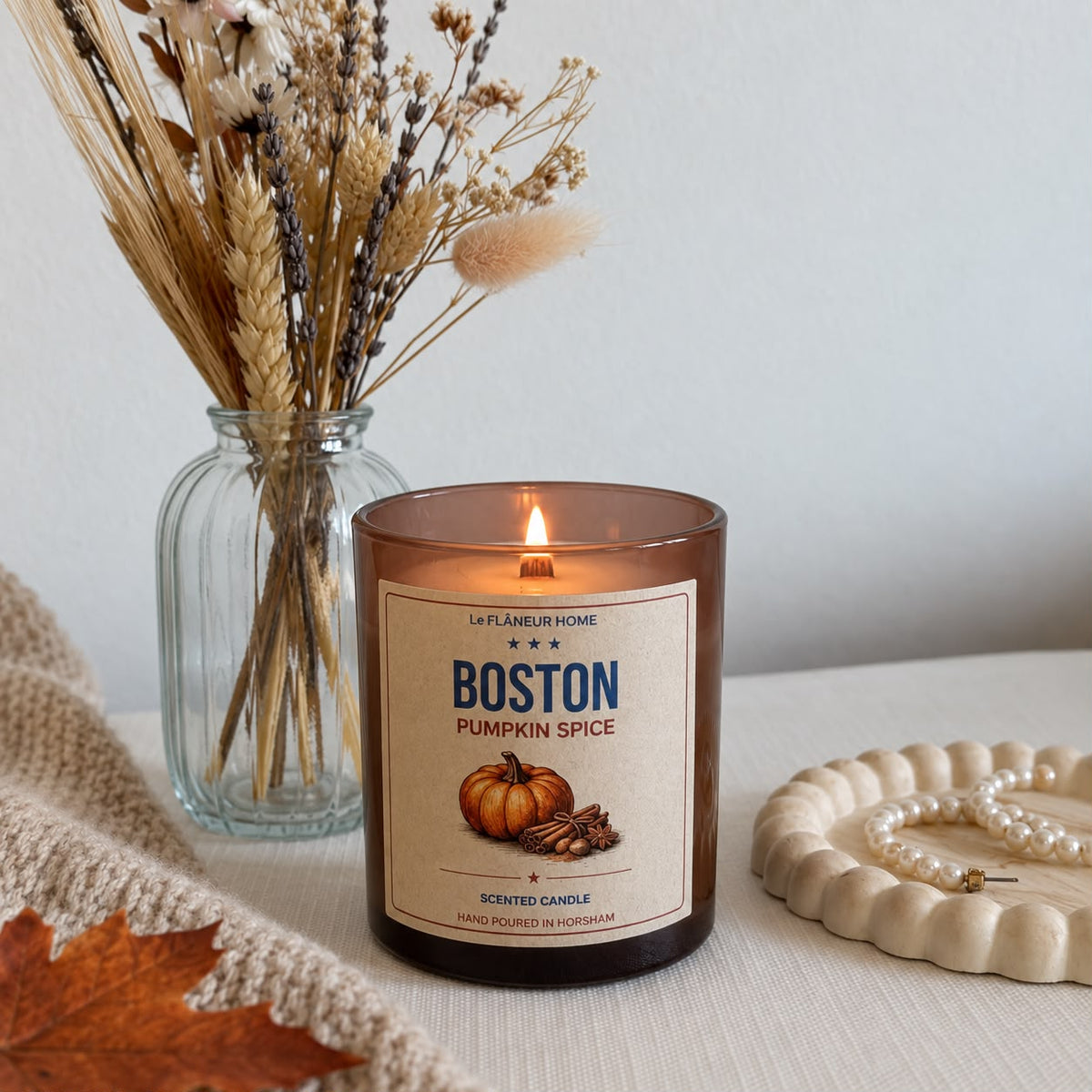 Lit Boston Pumpkin Spice candle in amber glass beside dried flowers, a knitted throw and a pearl necklace.