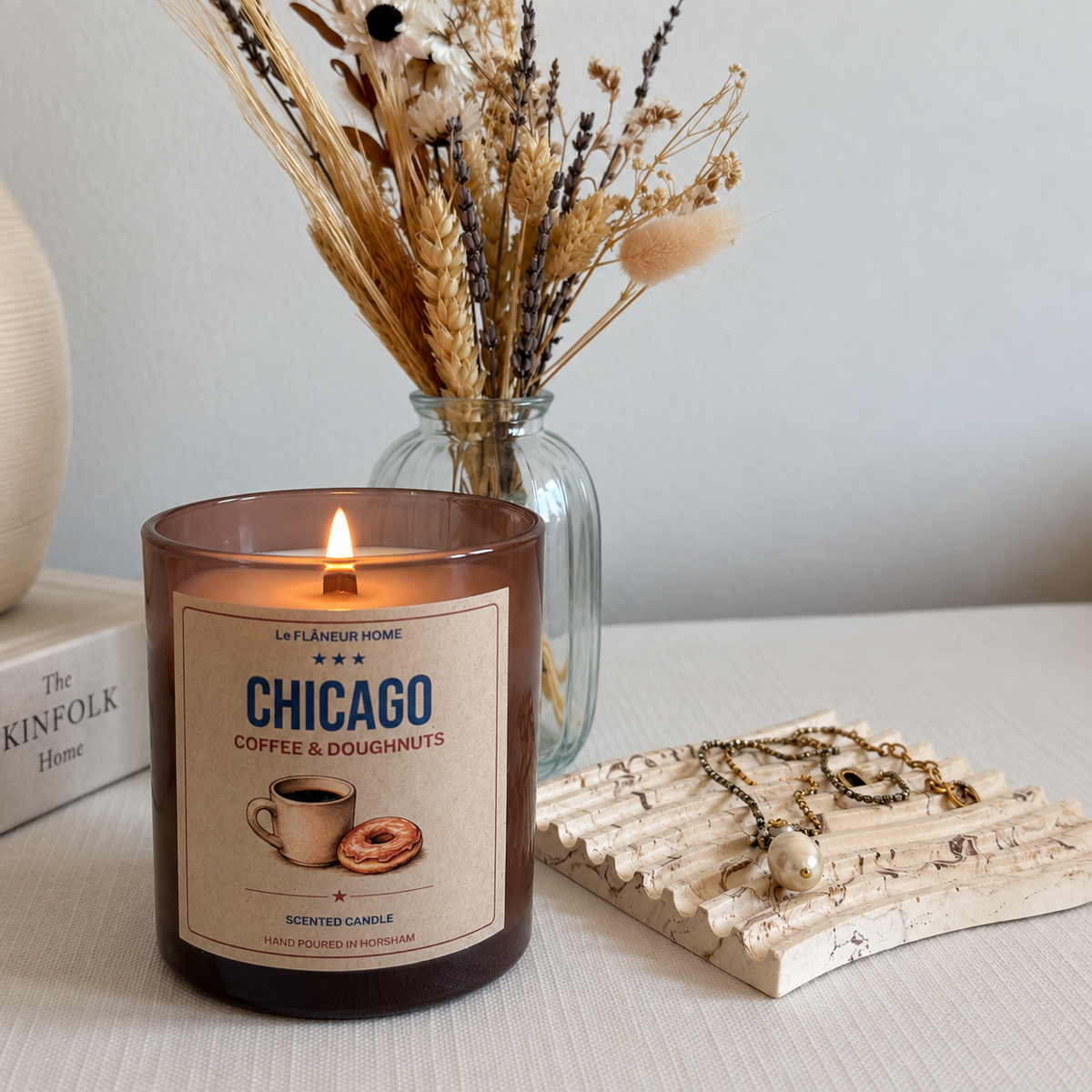 Lit Chicago Coffee & Doughnuts candle in amber glass beside dried flowers and jewellery on a cream tabletop.