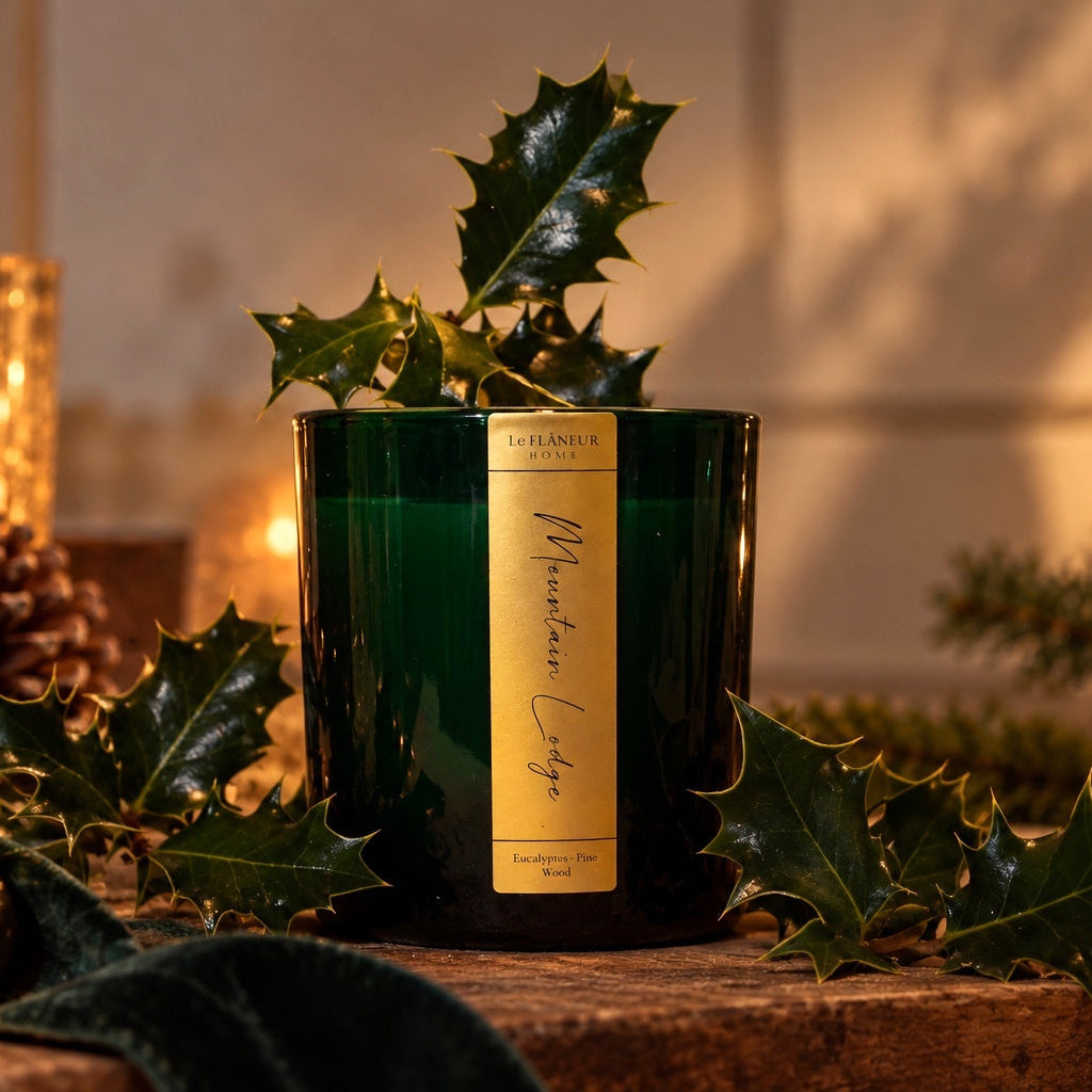 Le Flâneur Home Mountain Lodge scented candle in green glass with a gold label, styled with holly and pine cones.