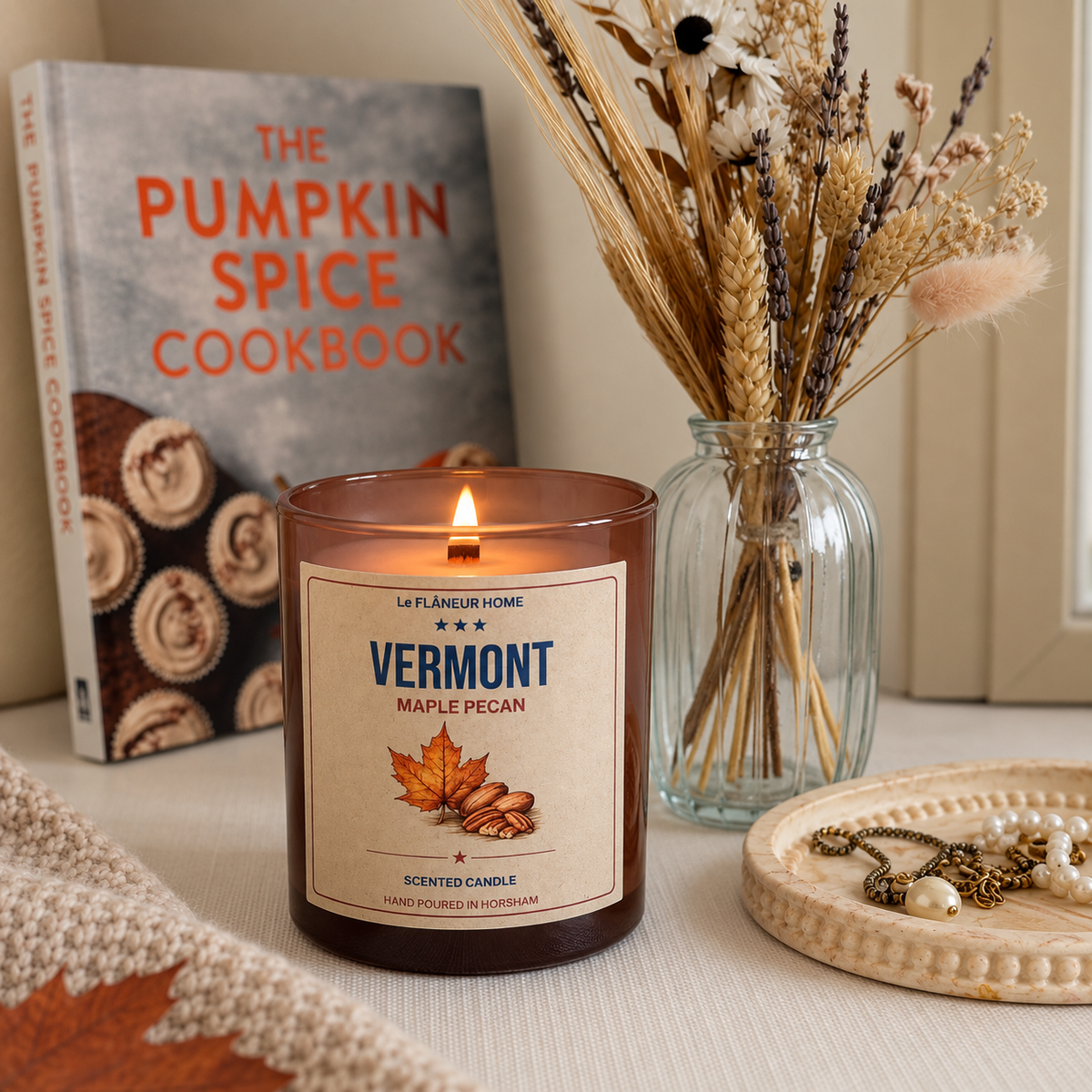 Lit Vermont Maple Pecan candle in amber glass beside dried flowers and a cookbook on a cream tabletop.